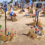 A person in uniform stands amid a memorial with photos, flowers, and flags attached to poles in dry, open ground on a sunny day.
