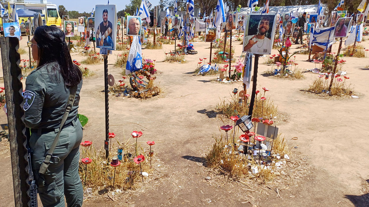 A person in uniform stands amid a memorial with photos, flowers, and flags attached to poles in dry, open ground on a sunny day.