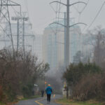 Two people walk on a paved trail surrounded by bushes and trees, with power lines stretching overhead and the tall buildings of Dominion visible through the mist in the background.