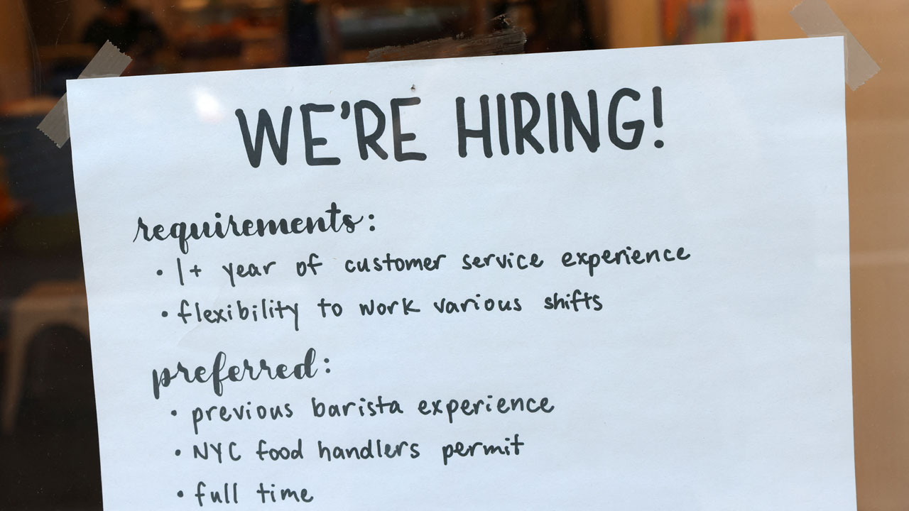 A sign on a window reads: WERE HIRING! Requirements: 1+ year of customer service, flexibility for shifts. Preferred: barista experience, NYC food handlers permit, full time..