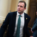 Max Miller wears a dark suit, light dress shirt, and patterned green tie while walking through a government building. An American flag and another man in a suit are visible in the blurred background.
