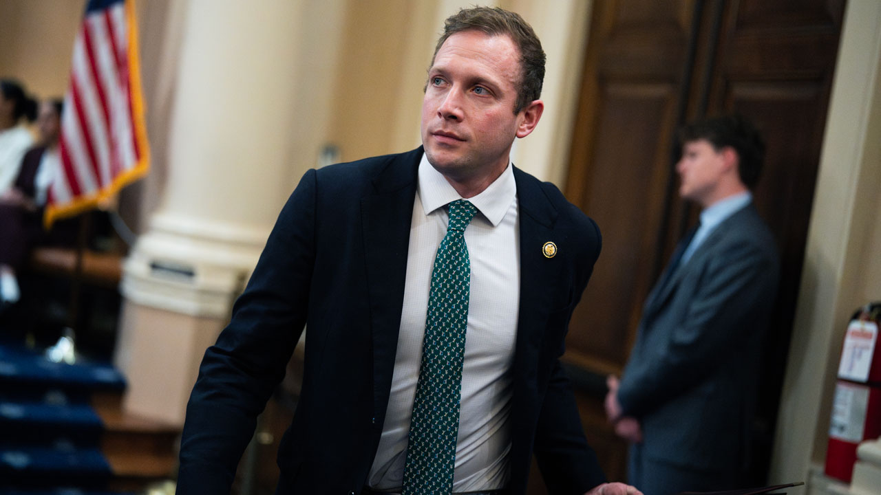 Max Miller wears a dark suit, light dress shirt, and patterned green tie while walking through a government building. An American flag and another man in a suit are visible in the blurred background.