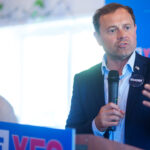 Tom Perriello speaks into a microphone at a podium with VOTE YES signs, while Hakeem Jeffries stands in the background.