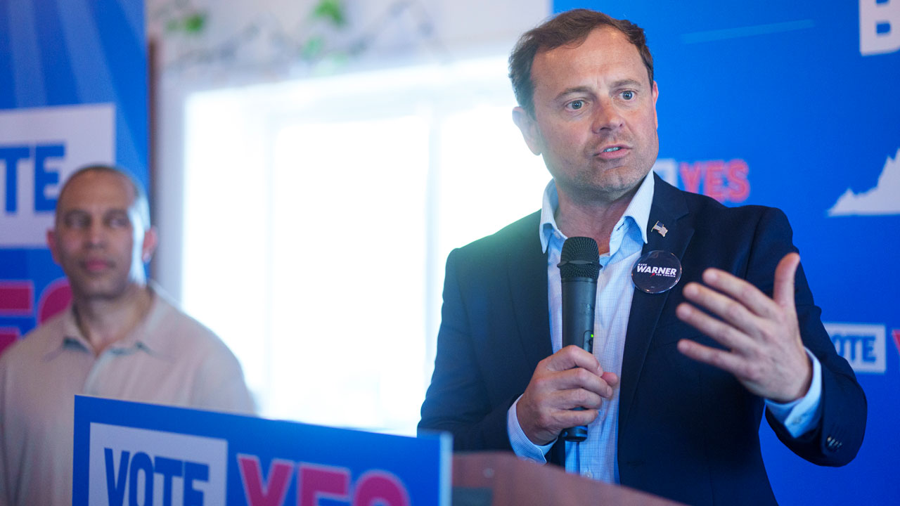 Tom Perriello speaks into a microphone at a podium with VOTE YES signs, while Hakeem Jeffries stands in the background.