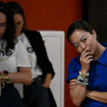 Francesca Hong, dressed in a blue shirt, stands with her arms crossed and hand near her mouth, looking thoughtful. Two people in white shirts stand in the background—one looking down and the other adjusting something.