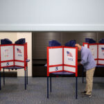 A man stands and fills out a ballot at a voting booth inside a polling station, with several booths visible beside him.