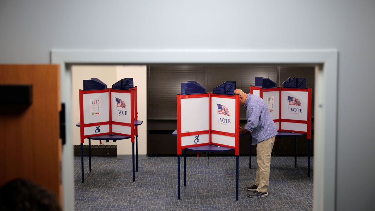 A man stands and fills out a ballot at a voting booth inside a polling station, with several booths visible beside him.