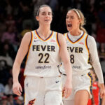 Caitlin Clark and Sophie Cunningham in white jerseys, Clark smiling and Cunningham shouting, walk side by side on a basketball court with a crowd and opposing players in the background.