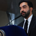 Zohran Mamdani in a suit speaks at a podium with a seal, using a microphone. He has dark hair and a beard, and appears to be addressing an audience indoors.