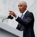 Barack Obama in a dark suit and tie, speaks at a gray podium with microphones, gesturing with his right hand against a light stone background.