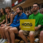 A group of people sit closely in a gym, watching an event. One person in front holds a yellow Francesca Hong for Governor sign. The crowd appears focused and attentive. Banners and a scoreboard hang in the background.