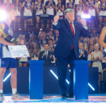 President Donald Trump dances on stage with young athletes holding trophies and checks at the Patriot Games. A cheering audience is in the background, making the scene appear festive and celebratory.