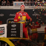NASCAR race car driver Joey Logano in a yellow Pennzoil suit celebrates atop his car in victory lane as confetti falls. Crew members behind him cheer and clap, while racing sponsor logos cover the background.