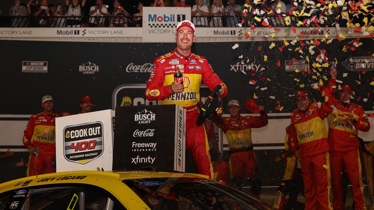 Home 1 NASCAR race car driver Joey Logano in a yellow Pennzoil suit celebrates atop his car in victory lane as confetti falls. Crew members behind him cheer and clap, while racing sponsor logos cover the background.