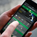 A person uses a smartphone to access a sports betting app, showing various sports icons and betting options on the screen.