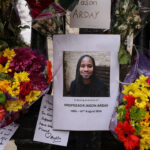 A memorial with colorful flowers surrounds a photo of a smiling man. The sign reads, In loving memory of Professor Jason Arday 1985–14th August 2026. Messages and bouquets are arranged around the tribute.