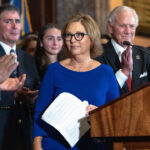 Darline Graham in a blue dress holding papers stands at a podium as people around her applaud. Several people in suits and formal attire are visible, and a flag is in the background.