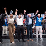 Abdul El-Sayed, Alexandria Ocasio-Cortez, Bernie Sanders and others stand on stage holding hands and raising them in unity; a cheering crowd with signs is behind them.