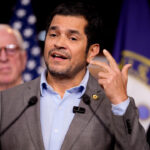 Rep. Jimmy Gomez, in a suit with no tie, speaks at a podium with two other men behind him. U.S. flags and an official emblem are visible in the background.