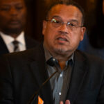 Keith Ellison glasses and a dark blazer speaks at a podium, with two people standing behind him, during an event in Minnesota.