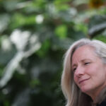 Abigail Spanberger with long gray hair smiles softly, sitting outdoors in Virginia with blurred green foliage and sunflowers in the background.