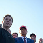 Elon Musk in a black shirt gestures while speaking to Donald Trump and a group of people outdoors.