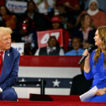 Donald Trump in blue suit and red tie sits on stage beside Sarah Huckabee Sanders in a blue dress holding a microphone, with several women visible among the crowd and patriotic banners in the background.