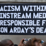 A black sign with white lettering holds "Racism within mainstream media is responsible for Jason Arday's death."