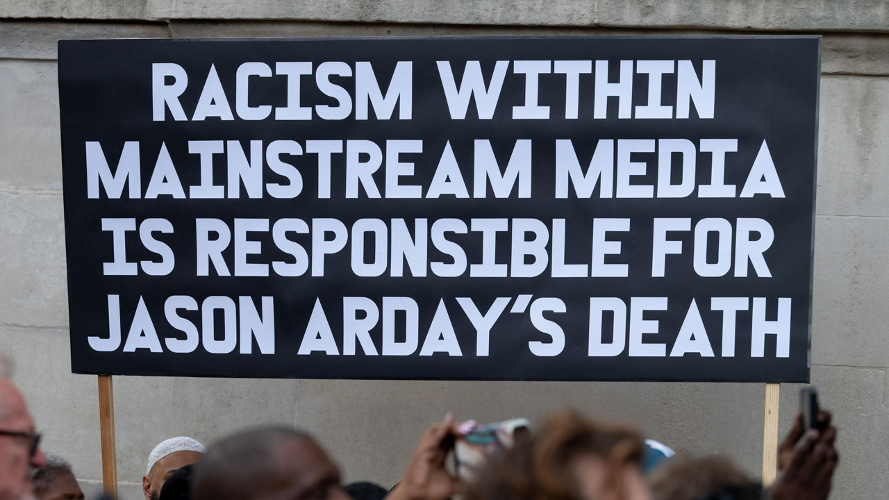 Home 1 A black sign with white lettering holds "Racism within mainstream media is responsible for Jason Arday's death."