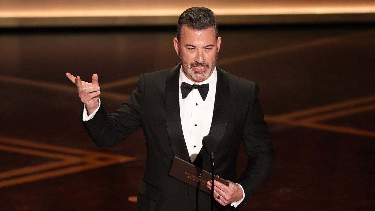 A man in a black tuxedo stands on stage, holding an envelope and gesturing with one hand while speaking into a microphone at a formal event.