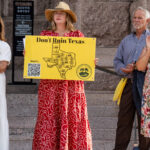 Four adults stand on stone steps at a rally. One woman in a red dress and hat holds a yellow sign reading “Don’t Ruin Texas,” protesting concerns over the new Data Center project. The others wear summer clothes, and a stone building is visible in the background.