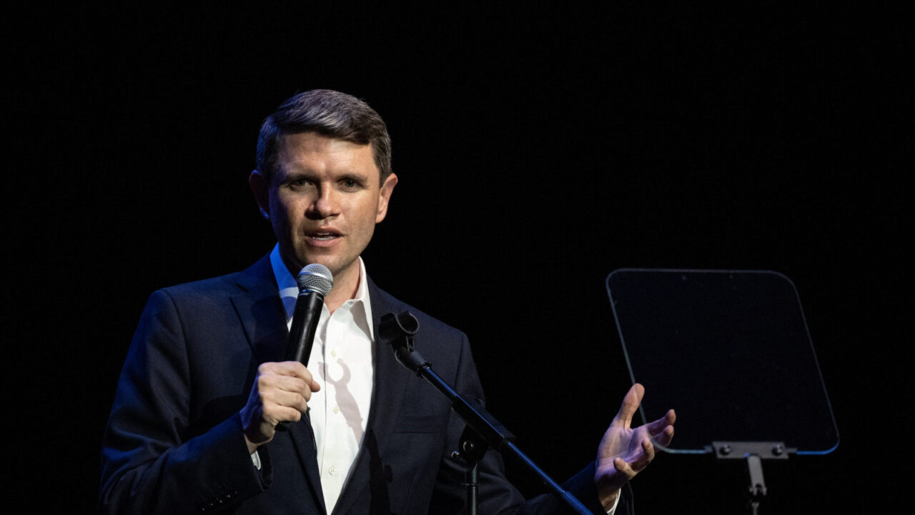 James Talarico in a suit speaks into a microphone on stage, gesturing with one hand, with a transparent teleprompter next to him against a dark background.