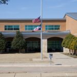 A brick school building with two flags at half-staff in front, surrounded by trimmed trees and a sidewalk under a clear blue sky.