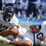 Two football players in white Virginia uniforms are on the field; one is holding the ball while laying down, and the other is behind him, with a blurred crowd and players in blue in the background.