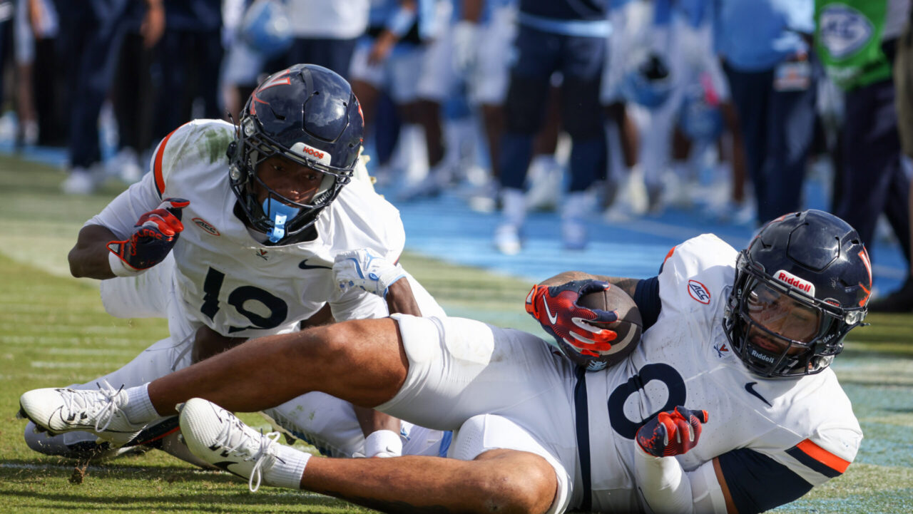 Two football players in white Virginia uniforms are on the field; one is holding the ball while laying down, and the other is behind him, with a blurred crowd and players in blue in the background.