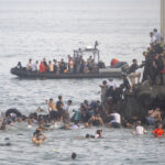 A large group of people in the water near a rocky shore in Spain, with some climbing rocks and others watched by uniformed officers; boats and more people are seen in the background.