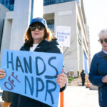 Protesters hold signs saying HANDS OFF NPR and Defend PBS & NPR outside a building during a daytime rally.
