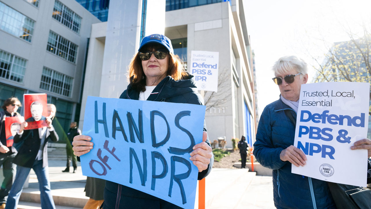 Home 4 Protesters hold signs saying HANDS OFF NPR and Defend PBS & NPR outside a building during a daytime rally.