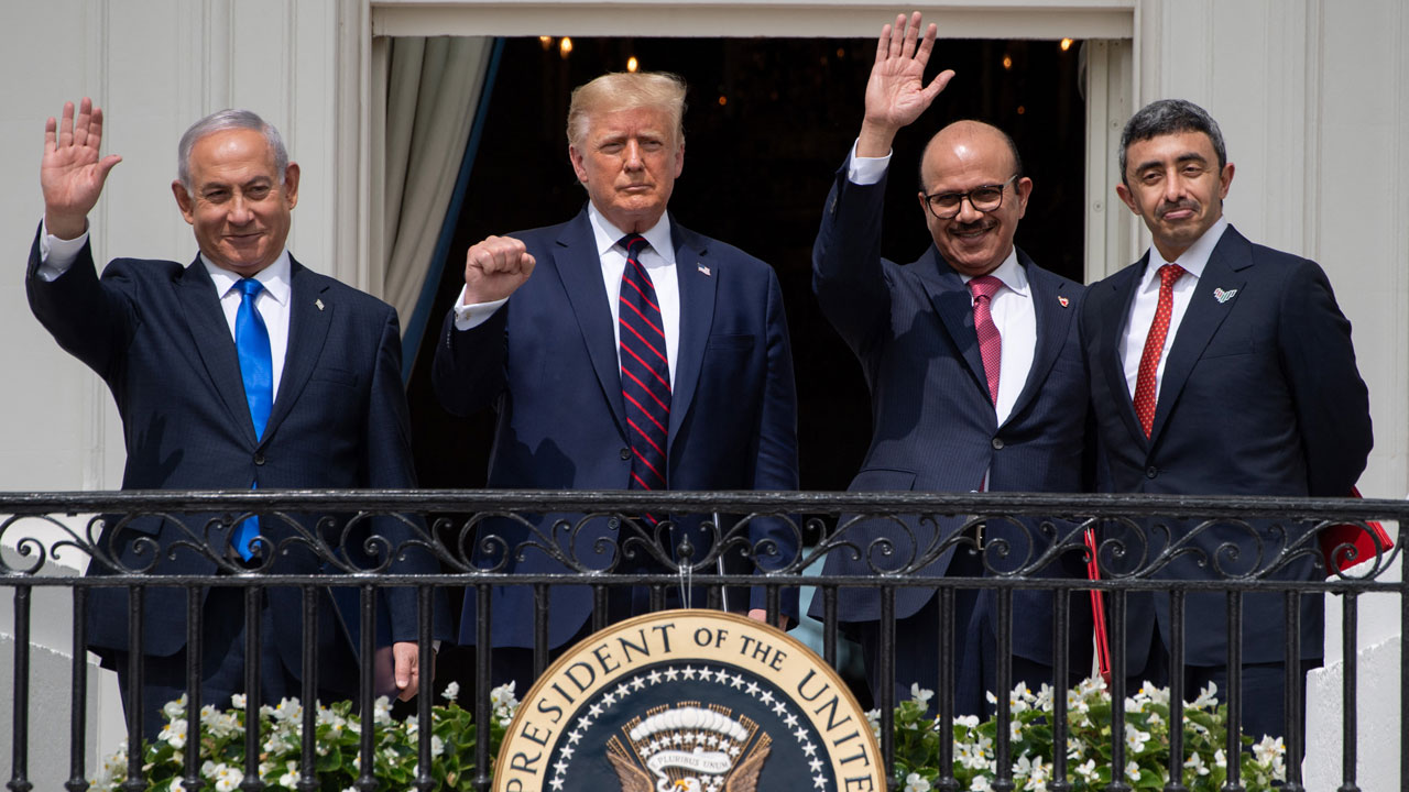 Four men in suits stand on a balcony behind the U.S. presidential seal, waving and smiling, with a white curtain and window in the background.