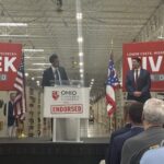Republican nominee for Ohio governor, Vivek Ramaswamy, looks on at his running mate, Senate President Rob McColley as they stand on a stage in a warehouse, with Vivek for Ohio campaign signs and American and Ohio flags behind them.