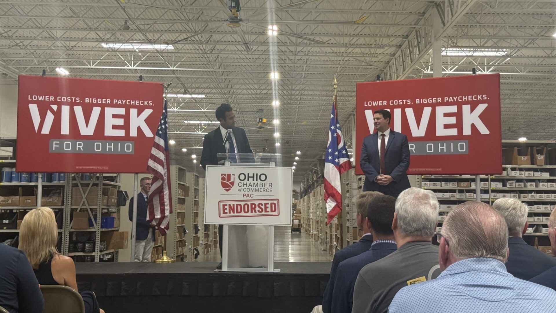 Republican nominee for Ohio governor, Vivek Ramaswamy, looks on at his running mate, Senate President Rob McColley as they stand on a stage in a warehouse, with Vivek for Ohio campaign signs and American and Ohio flags behind them.