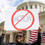 A woman stands outside of the Ohio capitol with a prohibition slash through symbol sign of "Acton."
