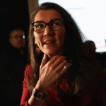 A woman wearing glasses and a maroon outfit stands indoors, touching her neck and smiling, with several people and a large bright screen in the background.