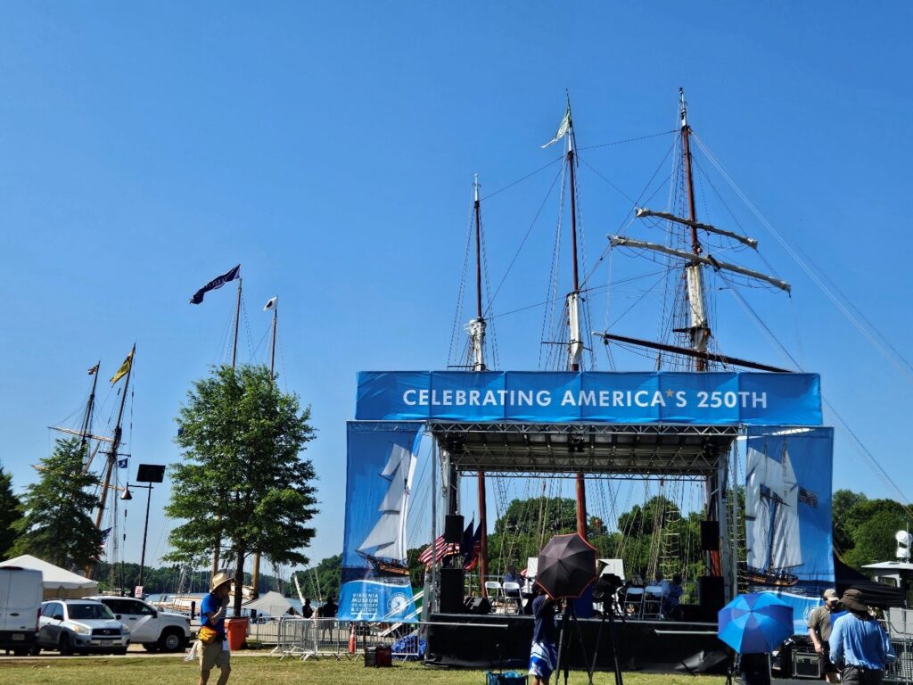 Tall Ships Bring the Spirit of ‘76 to Virginia 2 An outdoor stage with banner "Celebrating America's 250" with the masts of three tall ships rising in the background