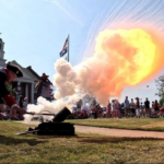A person in historical attire fires a cannon, creating a bright explosion and smoke, as a crowd watches outdoors near a historic South Carolina building with a flag in the background.