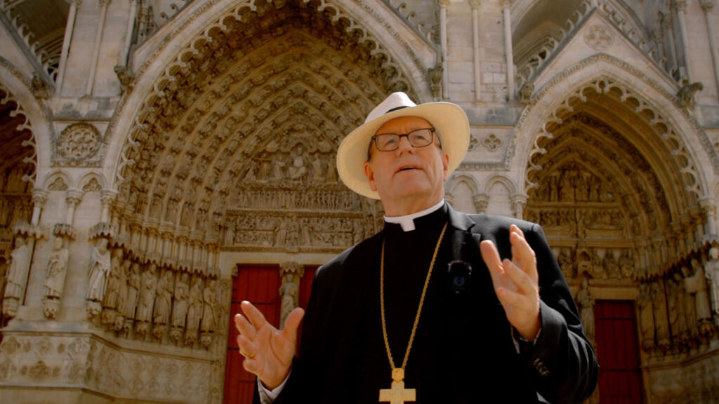 A clergyman in a white hat and black robe with a cross necklace gestures while standing in front of an ornate, arched cathedral entrance adorned with detailed carvings.