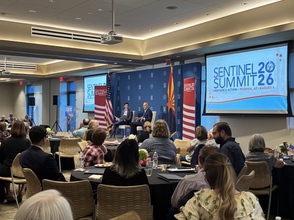 Arizona Rep. Andy Biggs Gives Grassroots Leaders Optimistic Inside Look at 'Battle' for America’s Future 1 A conference room filled with attendees seated at tables, watching a panel of speakers on stage—including Rep. Andy Biggs. Large screens display Sentinel Summit 2026 along with event details. American flags and banners decorate the space.
