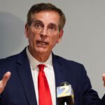 A man in a navy suit, red tie, and glasses speaks at a podium, gesturing with both hands raised. A microphone with a yellow and blue 2 logo is visible in front of him against a plain background.