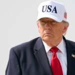 President Donald Trump wears a dark suit, red tie, and a white USA hat standing outside against a plain, light background.