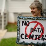 A smiling older woman holds a handmade sign outlined with tinsel that reads No Data Centers, with Data Centers crossed out by a red circle and slash. She stands outdoors in front of a light-colored building.